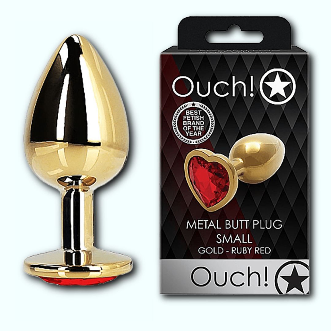 A Shots Ouch! Heart Gem Butt Plug Small in gold with a ruby red gem base is displayed next to its packaging. The box, branded "Shots," showcases the product and describes it as having a small, tapered tip in gold and ruby red hues. It also features a "Best Fetish Brand of the Year" badge.