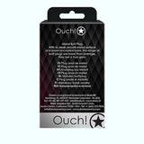 A retail package for the "Shots Ouch! Heart Gem Butt Plug Small - Gold/Ruby Red" features a sleek black design adorned with a star logo, the product name, and a multi-language description. The text emphasizes its smooth metal surface, jewel-encased base, and tapered tip for comfortable use during temperature play.