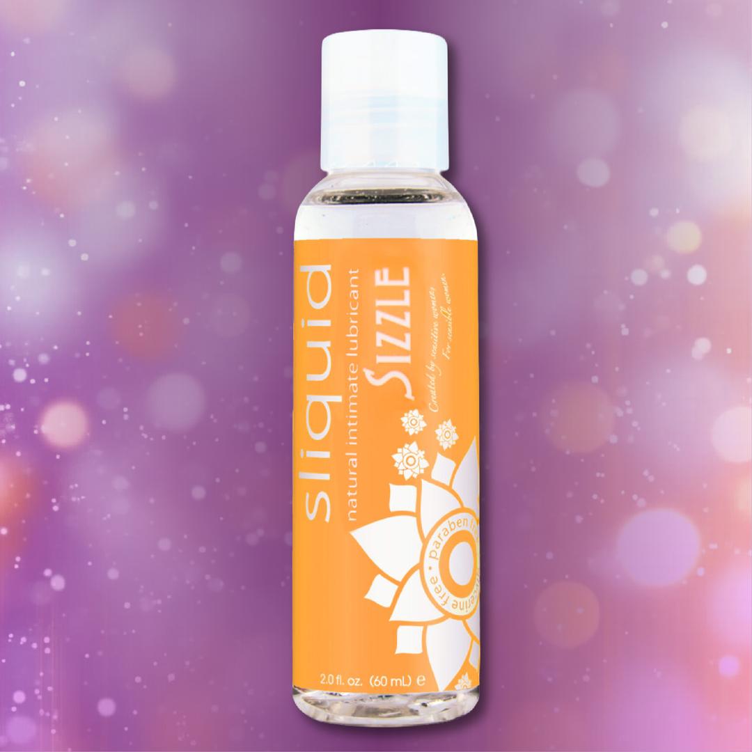 A 2 oz bottle of Sliquid Naturals ‘Sizzle’ Warming Lubricant by Sliquid, featuring an orange label, is set against a softly blurred purple background with bokeh light effects, highlighting the inviting appeal of this water-based warming lubricant.