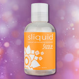 A clear bottle of Sliquid Naturals ‘Sizzle’ Warming Lubricant by Sliquid, featuring an orange label, set against a pink and purple bokeh background.