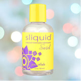 A bottle of Sliquid Naturals Swirl Pina Colada Flavored Lube, free of glycerin and parabens, features a yellow and purple floral label. This 4.2oz water-based lubricant is elegantly displayed against a soft bokeh effect in pastel colors, perfectly balancing design and functionality.