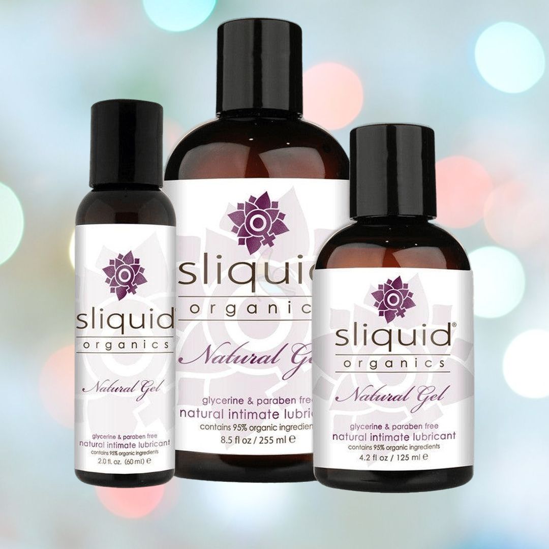 Three bottles of Sliquid Organics Natural Ultra Thick Aloe-Based Lube, which features organic botanical extracts, are elegantly presented with their sleek black bottles and white labels prominently displaying the brand and product details. A softly blurred background in blue and pink tones enhances the presentation.