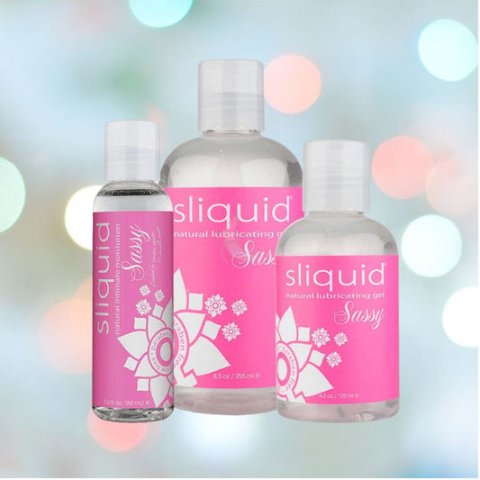 Three bottles of Sliquid Sassy Ultra-Thick Lubricant, ideal for use as an anal gel, in different sizes are showcased against a gentle, colorful bokeh backdrop. The bottles have pink labels decorated with floral motifs. 1080