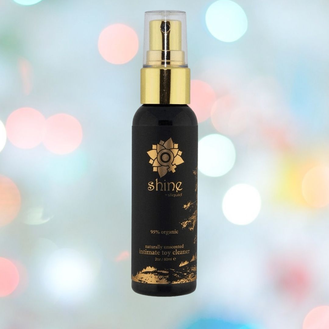 A bottle of Sliquid Shine Organic Sex Toy Cleaner in black and gold stands against blurred bokeh lights. This vegan, organic cleaner is unscented, contains 95% organic ingredients, and has antibacterial properties for safe, gentle care.