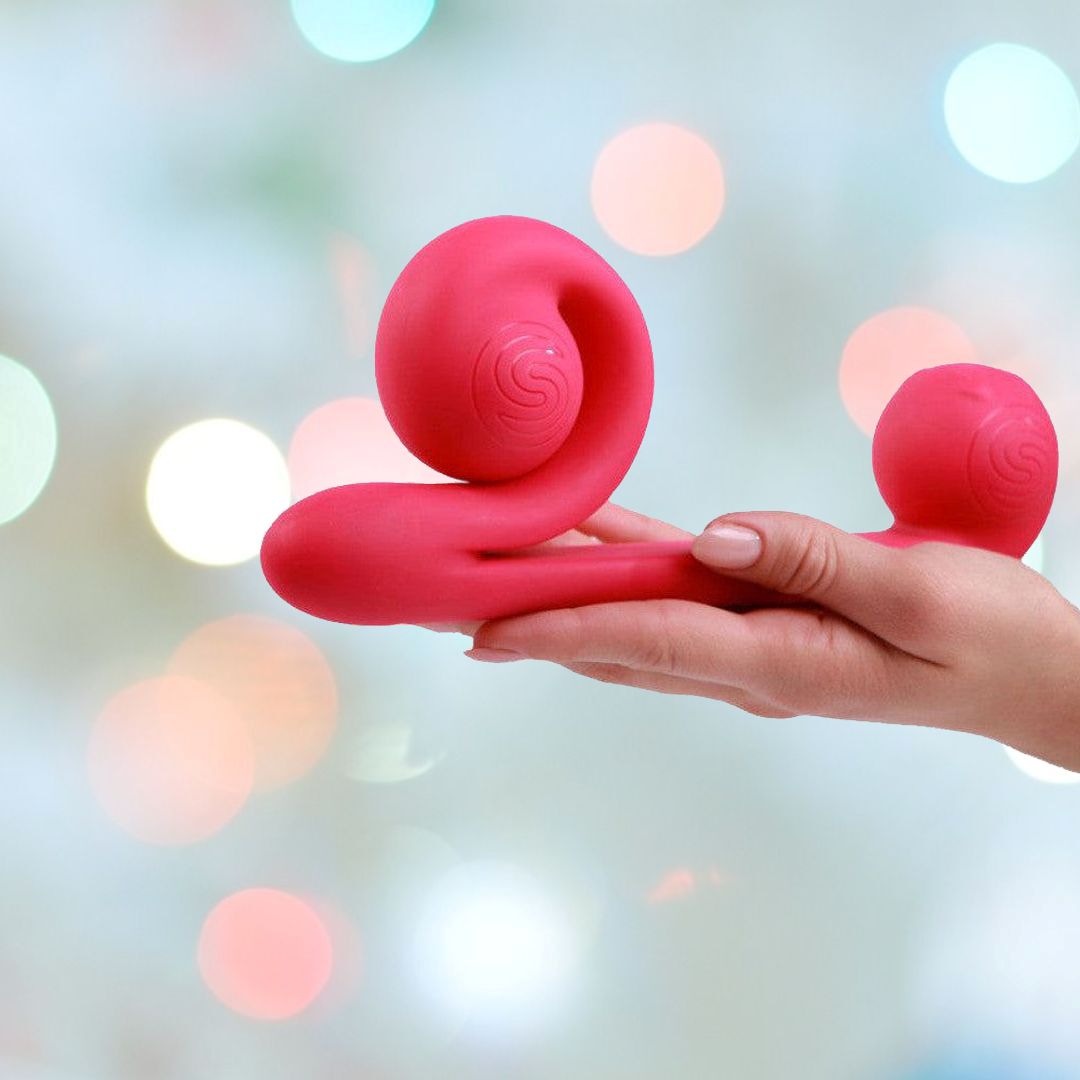 A person holds a pink Snail Vibe Dual-Motor Thrusting Vibrator with two rounded ends against a blurred background of soft, multicolored bokeh lights.