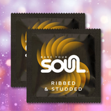 Two black packets of Soul Ribbed & Studded Latex Condoms, featuring a yellow swirl design and the brand name “soul,” sit against a pink and purple sparkly bokeh background. These premium latex condoms are expertly crafted by soul with ribbed and studded textures for enhanced pleasure.