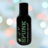 A black bottle labeled "SPUNK Pure Silicone Lubricant" by Spunk Lube is centered against a blurred background with colorful bokeh lights. The 2 fl. oz. (59 ml) bottle features a long-lasting slippery texture, ideal for extended use and heightened pleasure.