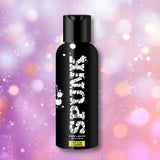 A black bottle labeled "SPUNK Hybrid Lubricant" from Spunk Lube, featuring a white splatter design, is centered against a blurred pastel background with soft bokeh effects.