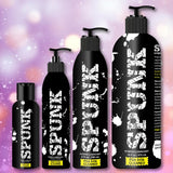 Four black bottles of Spunk Lube's SPUNK Hybrid Lubricant are lined up against a soft, colorful bokeh background. The largest has a pump dispenser. Each label reads “FDA 510K CLEARED.”.