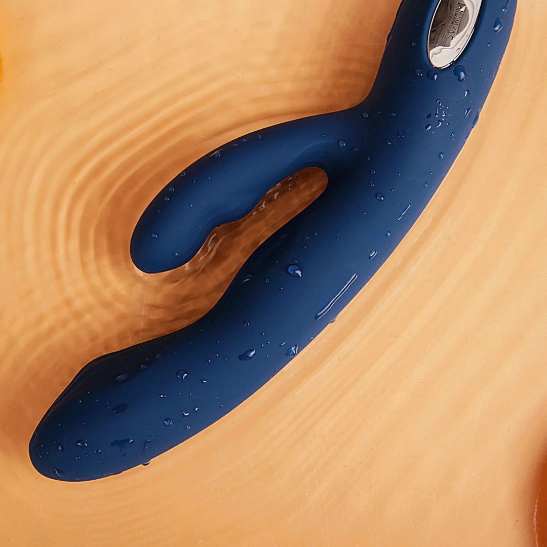 A Svakom Aylin Rabbit Vibrator in dark blue, featuring a curved design and pulsating G-spot tip, rests on a rippled light brown surface with water droplets highlighting its various vibration modes.