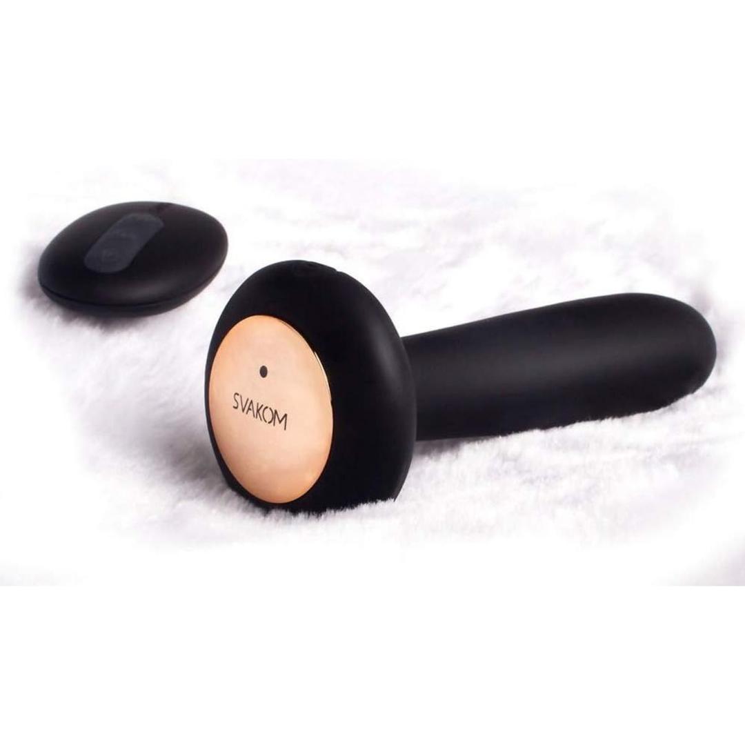 The Svakom Primo Heating Butt Plug, a sleek black cylindrical massager with gold accents, lies on a white fluffy surface alongside its oval remote, highlighting its convenient remote control and heating features.