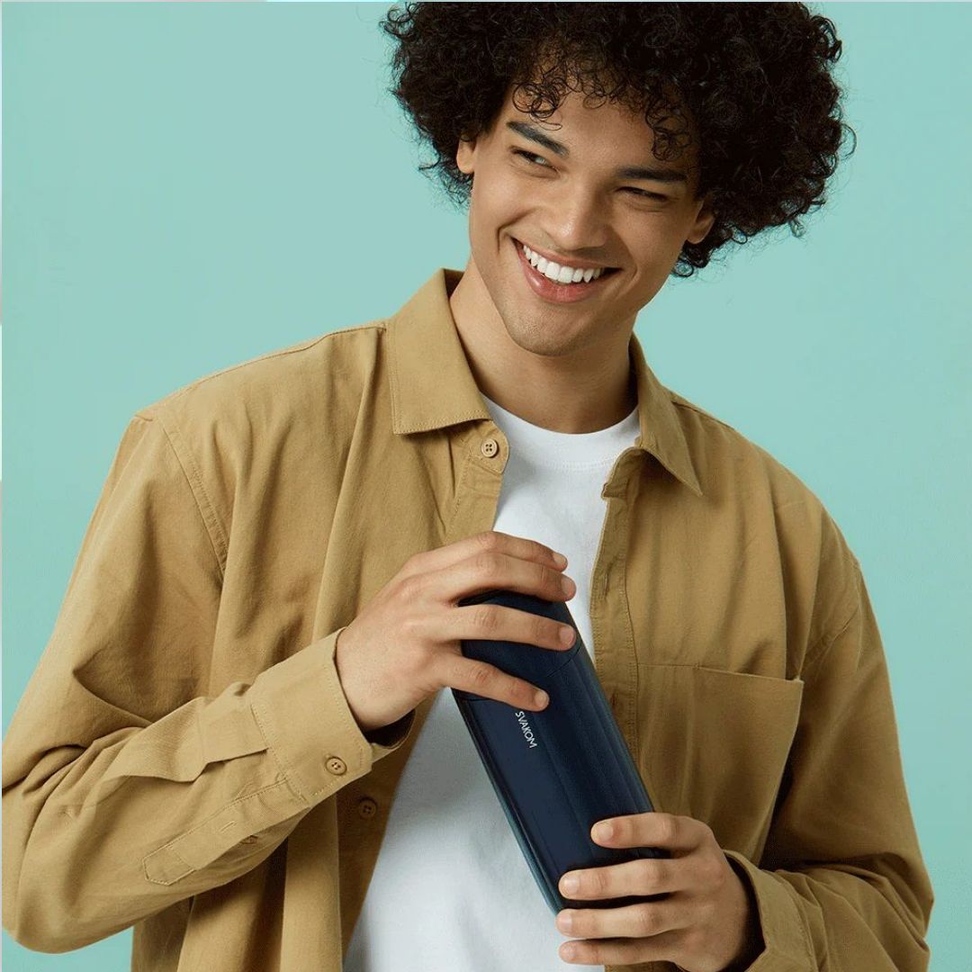 A person with curly hair, wearing a beige shirt, smiles while holding a dark blue bottle against a light teal background, enjoying the customizable experience of the Svakom Sam Neo 2 Sucking and Vibrating Masturbator by svakom.