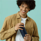 A person with curly hair, wearing a beige shirt, smiles while holding a dark blue bottle against a light teal background, enjoying the customizable experience of the Svakom Sam Neo 2 Sucking and Vibrating Masturbator by svakom.