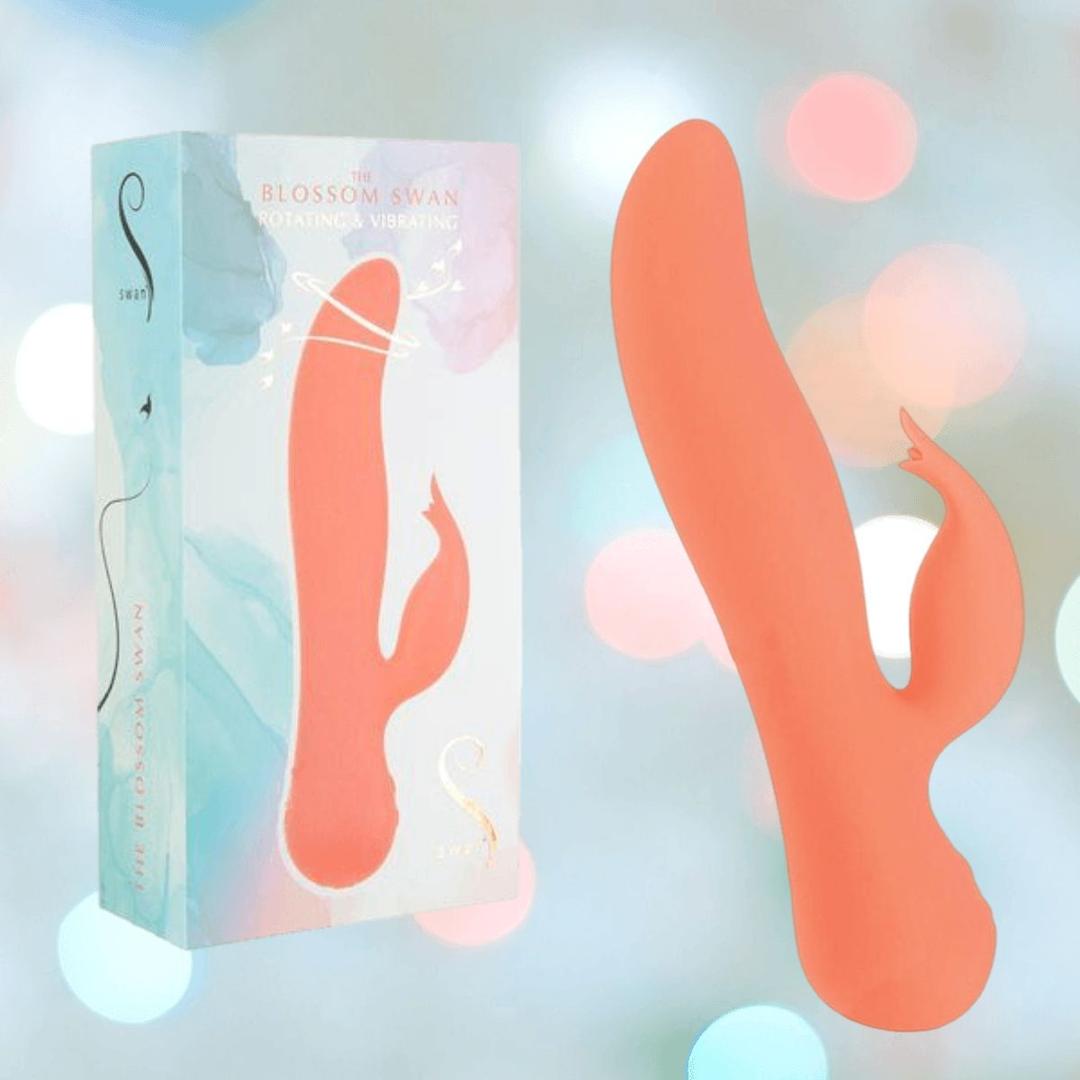 The Swan The Blossom Rabbit Vibrator - Coral by BMS, featuring a unique curved orange silicone design, is displayed beside its pastel box. The softly blurred background highlights the product.