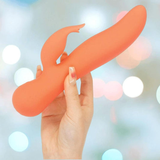 A hand with manicured nails holds the BMS Swan The Blossom Rabbit Vibrator in Coral, featuring a curved design and a smaller attachment, set against a softly blurred, colorful background. 1080