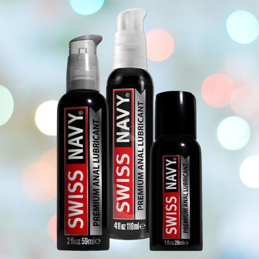 Three bottles of Swiss Navy Premium Anal Lubricant with Clove stand against a blurred background of colorful lights. The black bottles with red and white labels come in three sizes: 1 oz (29 ml), 2 oz (59 ml), and 4 oz (118 ml). Experience their long-lasting formula for unparalleled comfort. 1080