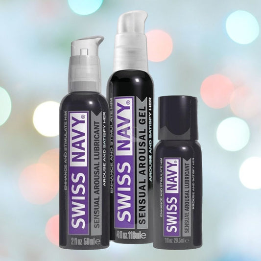 Three bottles of Swiss Navy Arousal Lubricant are arranged in front of a blurred background adorned with multicolored lights. These black bottles, showcasing purple and silver labels, emphasize their function in boosting natural lubrication. 1080