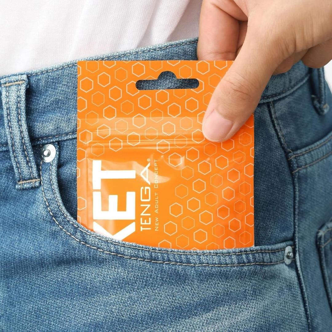 A hand places a Tenga Pocket Hexa Brick—a disposable textured male masturbator in a hexagon-patterned, orange resealable packet—into the front pocket of blue jeans. The Tenga brand name and some text are partially visible on the packaging.
