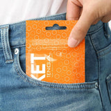 A hand places a Tenga Pocket Hexa Brick—a disposable textured male masturbator in a hexagon-patterned, orange resealable packet—into the front pocket of blue jeans. The Tenga brand name and some text are partially visible on the packaging.
