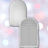 Two white, textured silicone scrubbers with small triangular bristles sit before a soft bokeh of pink, blue, and white lights. Like the Tenga POCKET Crystal Mist Disposable Micro-Nub Masturbator, they suggest delicate, cooling sensations.