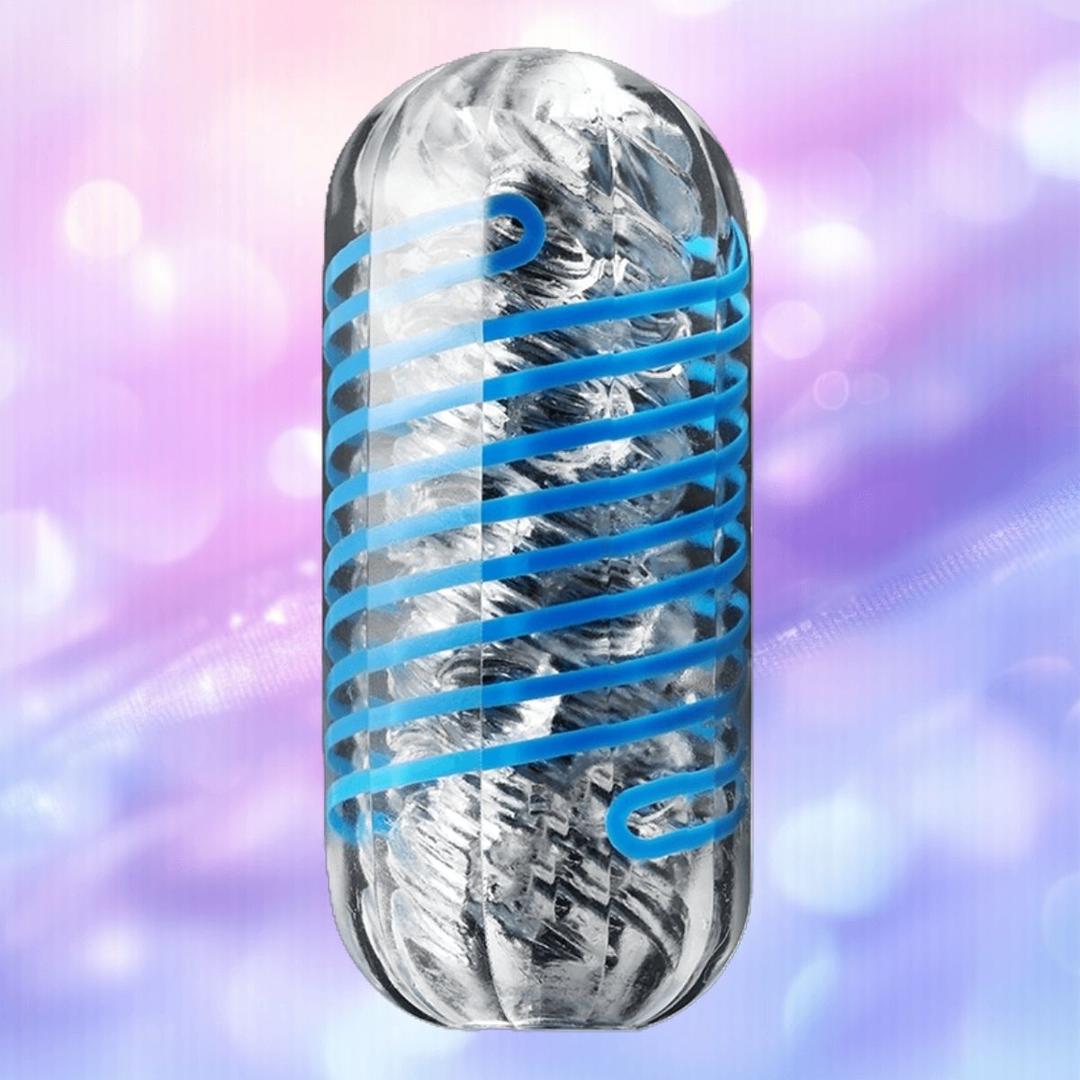 A silver metallic capsule with a blue spiral—resembling the Tenga Spinner DX 01 Drops by Tenga—set against a softly blurred pink and purple background with light bokeh effects.