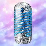 A silver metallic capsule with a blue spiral—resembling the Tenga Spinner DX 01 Drops by Tenga—set against a softly blurred pink and purple background with light bokeh effects.