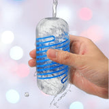 A hand holds the Tenga Spinner DX 01 Drops, a transparent reusable stroker masturbator with blue bands, under running water, with splashes and blurred white, pink, and blue lights in the background.