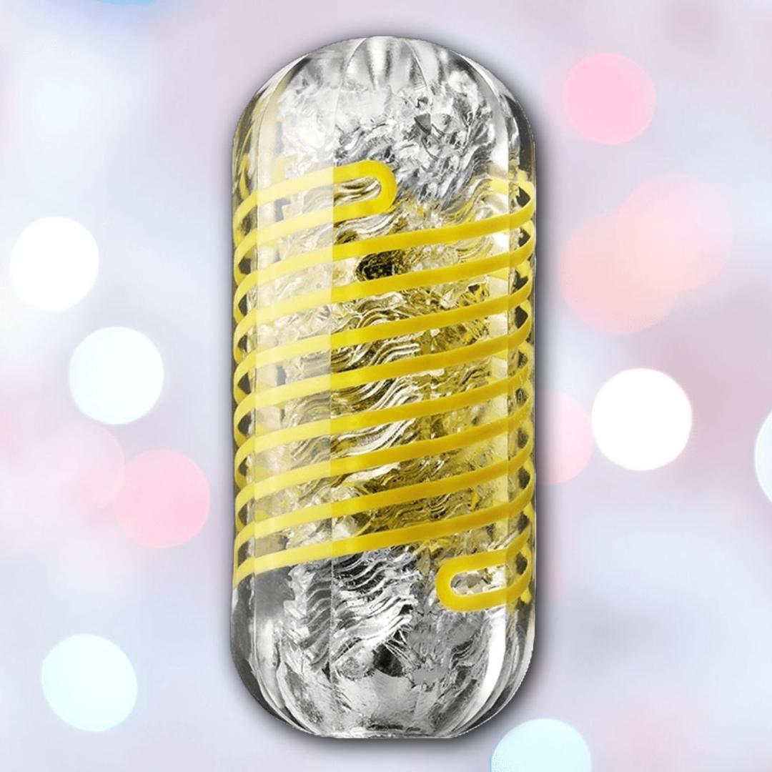 The Tenga Spinner DX 02 Bumps Reusable Stroker features a clear textured sleeve with a yellow spiral, offering dynamic spinning stimulation. Set against soft white and pink bokeh, this Tenga stroker stands out as a visually striking pleasure product.