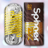 A transparent cylindrical Tenga Spinner DX 02 Bumps Reusable Stroker is shown with yellow spiral details and its brown-and-yellow packaging, set against a softly blurred pastel background.