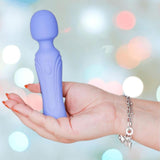A hand with painted nails and a silver bracelet holds The Swan Era Wand by BMS, which features incremental speed control and a PowerBullet motor, against a blurred background with pastel lights.
