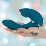 A hand with a floral tattoo holds the Swan The Monarch Swan - Teal vibrator by BMS against a softly blurred background with pastel-colored lights.