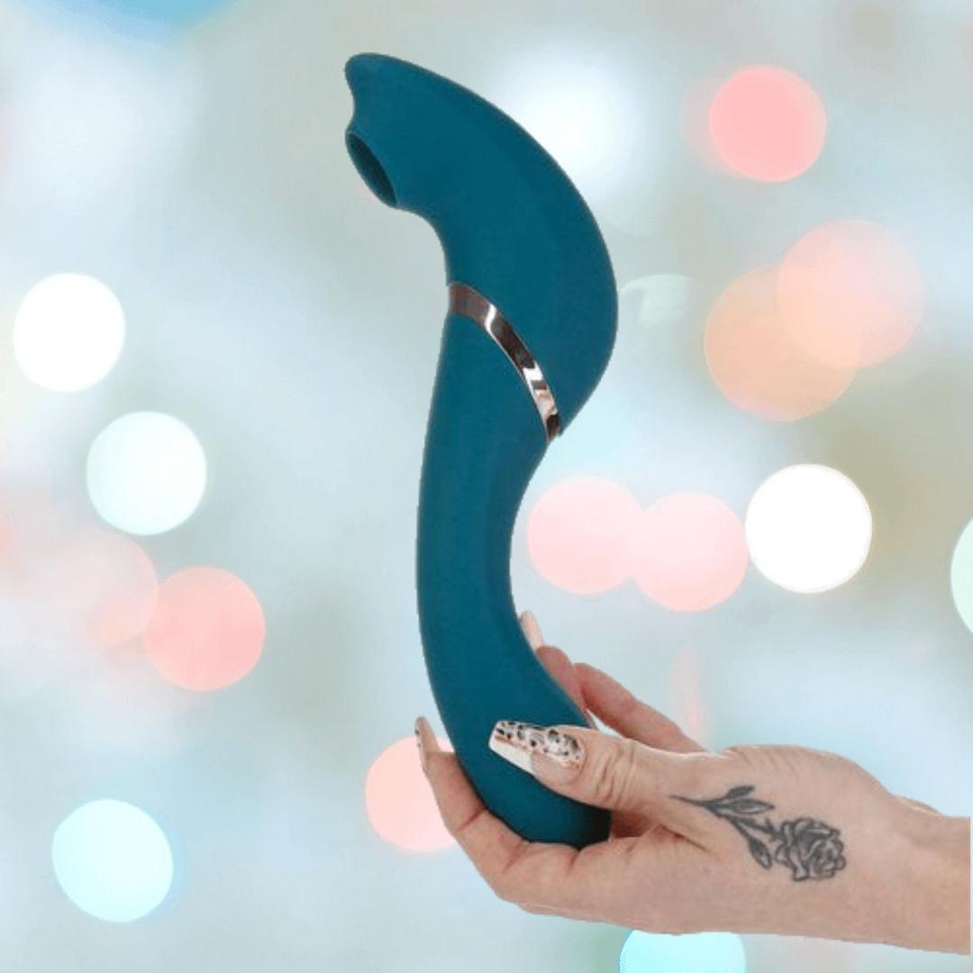 A hand with a floral thumb tattoo holds the luxurious BMS Swan The Monarch Swan vibrator in Teal, set against a soft background of colorful, blurred lights.