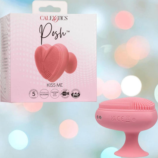 A pink heart-shaped CalExotics Posh Kiss Me Handheld Massager with textured pleasure nubbies and a suction base sits next to its branded box against a softly blurred pastel bokeh background. 1080