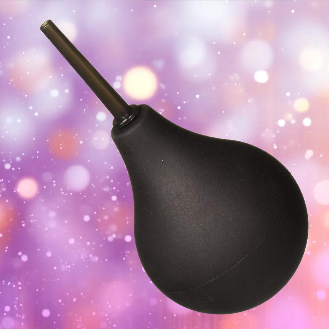 The Shibari Triton Easy Squeeze Enema Bulb, featuring a flexible tip and narrow nozzle, is shown at an angle against a blurred purple and pink background with circular light spots.