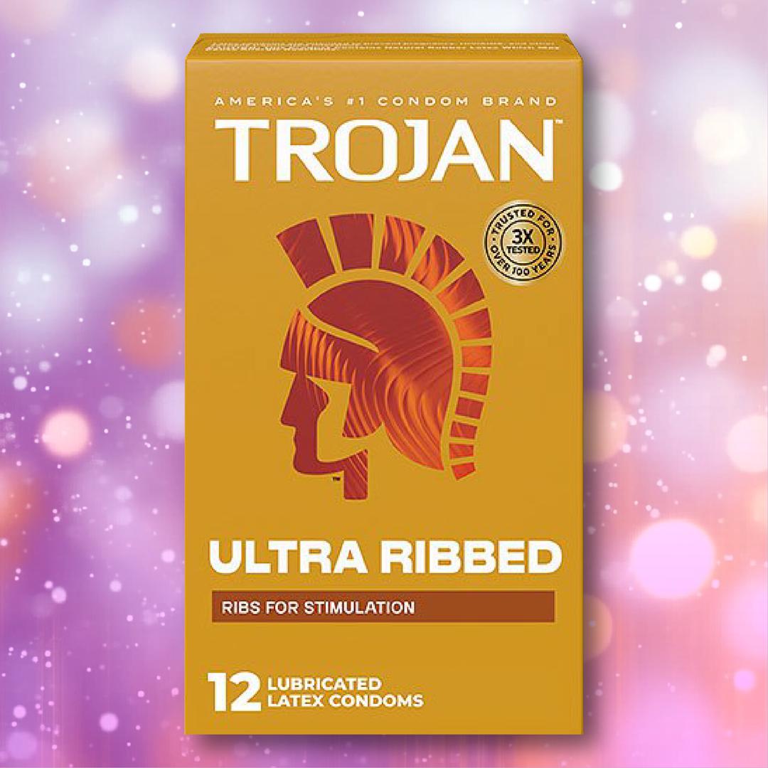 A box of Trojan Ultra Ribbed Condoms by Trojan, designed for added stimulation, featuring orange and yellow packaging with the helmet logo and “12 lubricated latex condoms” on a sparkly purple-pink background.