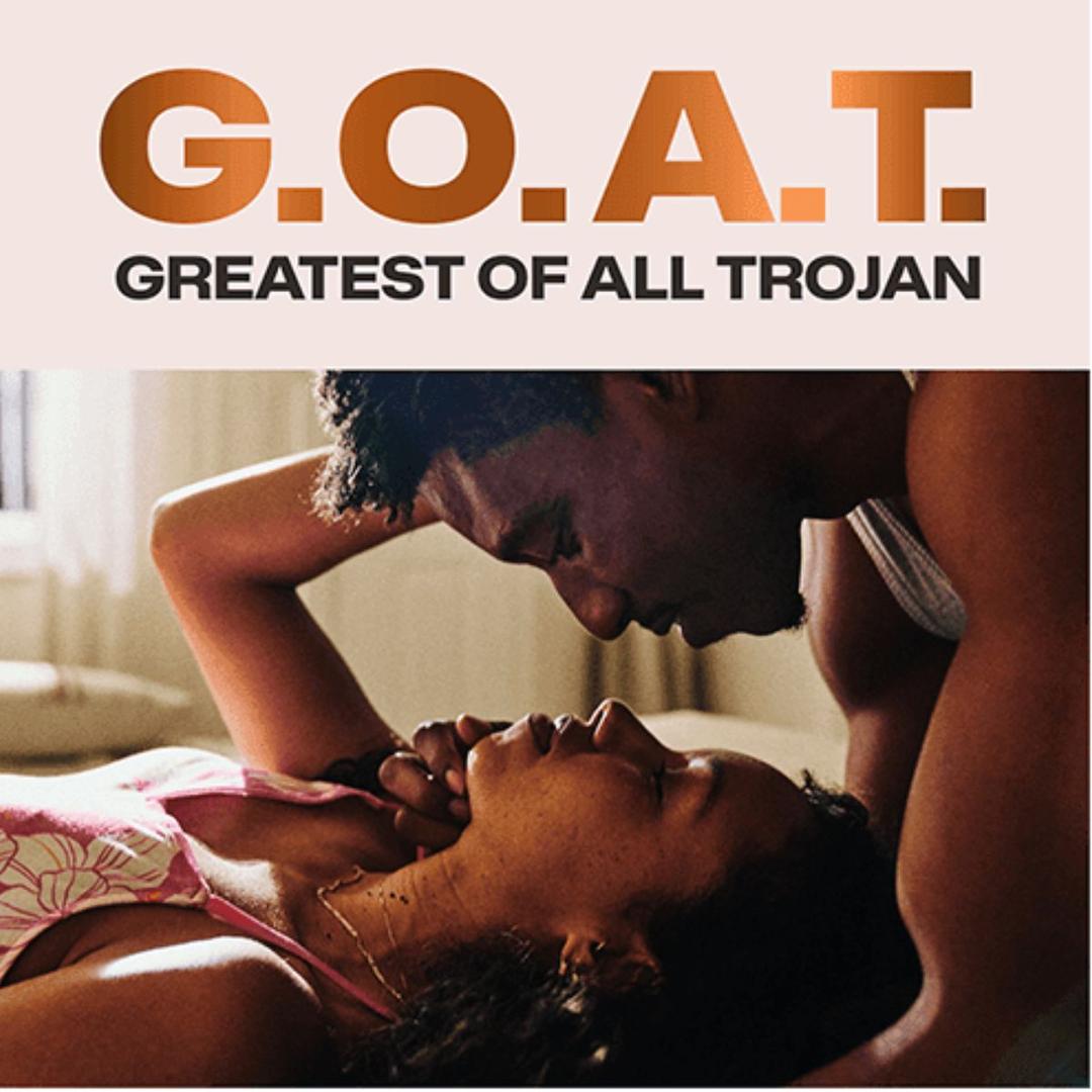 A couple lies close on a bed, gazing into each other's eyes. Bold text reads "G.O.A.T. Greatest Of All Trojan," spotlighting Trojan G.O.A.T. Non-Latex Lubricated Condoms for unforgettable comfort and connection.