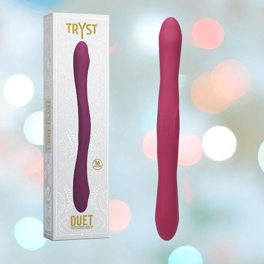 The Tryst Duet Double-Ended Silicone Vibrating Dildo with Wireless Remote by Doc Johnson in Berry is displayed next to its white decorative box, labeled "Tryst Duet," against a softly blurred pastel background.