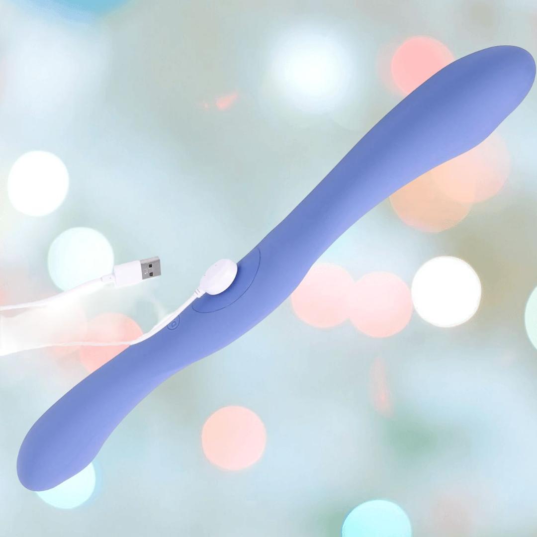 The Doc Johnson Tryst Duet Double-Ended Silicone Vibrating Dildo in periwinkle features two rounded ends and a wireless remote, shown with a USB charging cable against a soft, blurred pastel background.