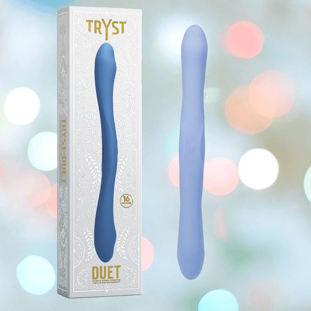 A periwinkle Tryst Duet Double-Ended Silicone Vibrating Dildo with Wireless Remote by Doc Johnson stands upright beside its white and gold packaging, set against a blurred, colorful background.