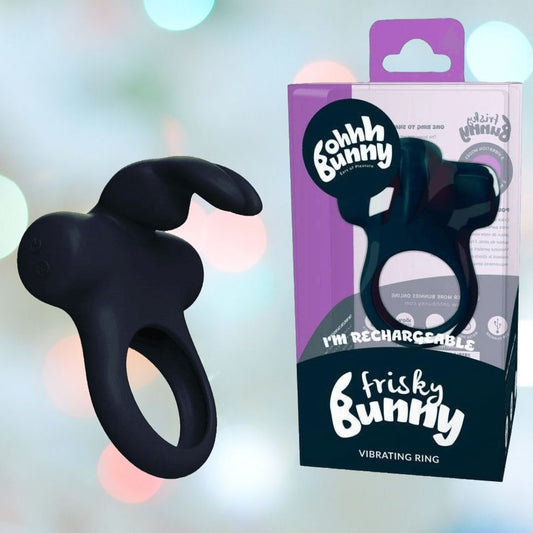 A VeDO Frisky Bunny Vibrating Cock Ring in black is positioned next to its packaging. The box is adorned in shades of purple and white, showcasing the brand "VeDO" along with the product name "VeDO Frisky Bunny Vibrating Cock Ring." The softly blurred background imbues a sense of mystery to this rechargeable vibrating ring. 1080