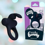 A VeDO Frisky Bunny Vibrating Cock Ring in black is positioned next to its packaging. The box is adorned in shades of purple and white, showcasing the brand "VeDO" along with the product name "VeDO Frisky Bunny Vibrating Cock Ring." The softly blurred background imbues a sense of mystery to this rechargeable vibrating ring.