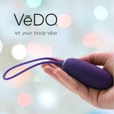A hand holds a purple VeDO Kiwi Bullet with a looped handle against a blurred, colorful background. The brand name "VeDO" and tagline "let your body vibe" appear in the top left corner.