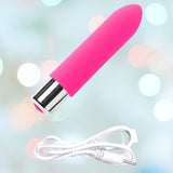 The VeDO Bam Mini Rechargeable Bullet Vibrator in pink, featuring a distinctive bullet shape and silver base, comes with a white USB charging cable. This sophisticated device stands gracefully against a blurred backdrop of soothing pastel hues.