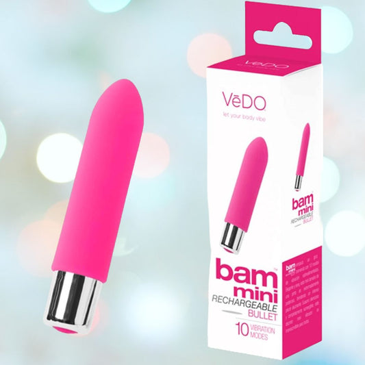 A VeDO Bam Mini Rechargeable Bullet Vibrator in pink, adorned with chrome details, is elegantly displayed next to its packaging that prominently features the "VeDO" brand name. The box lists features like "10 vibration modes," set against a softly blurred background of soothing pastel colors. 1080