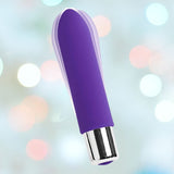 An indigo VeDO Bam Mini Rechargeable Bullet Vibrator, featuring a sleek design and silver base, is displayed against a soft, out-of-focus background with pastel bokeh lights. The background creates a dreamy, slightly festive atmosphere.