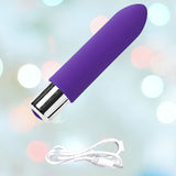 The VeDO Bam Mini Rechargeable Bullet Vibrator in Indigo, designed as a sleek cylindrical massager with a silver base, floats against a soft, blurred background with bokeh lights. Below the purple rechargeable bullet vibrator, a white USB charging cable is laid out neatly.