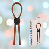 The Velv'Or Rooster Ragnar Adjustable Lasso String Cock Ring in brown is displayed with its packaging, featuring an image of the product. The background is softly blurred with pastel lights.