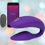 Introducing the We-Vibe Sync 2 Couples Vibrator with App in Purple, a device crafted by We-Vibe for shared pleasure. It boasts a curved design with an adjustable fit, accompanied by a remote control and app interface visible on a smartphone screen. The ambiance is set with shimmering blurred multicolored lights in the background, enhancing the enticing mood.