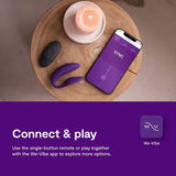 On a round wooden table sits a purple We-Vibe Sync 2 Couples Vibrator, alongside a small remote, a lit candle, and a smartphone open to the We-Vibe app. The text below says: "Connect & play. Experience shared pleasure with the couples vibrator using the one-button remote or join in together through the app.