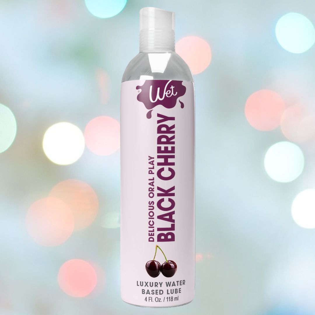 A transparent bottle of Wet Delicious Oral Play Black Cherry water-based flavored lubricant prominently displays the name "Delicious Oral Play Black Cherry" against a backdrop adorned with black cherry designs and soft, colorful bokeh lights.